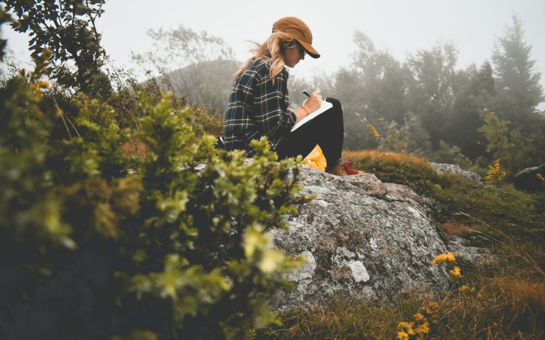 Person sitting outside in nature, writing intently in a journal, with a pen in hand and a calm expression.